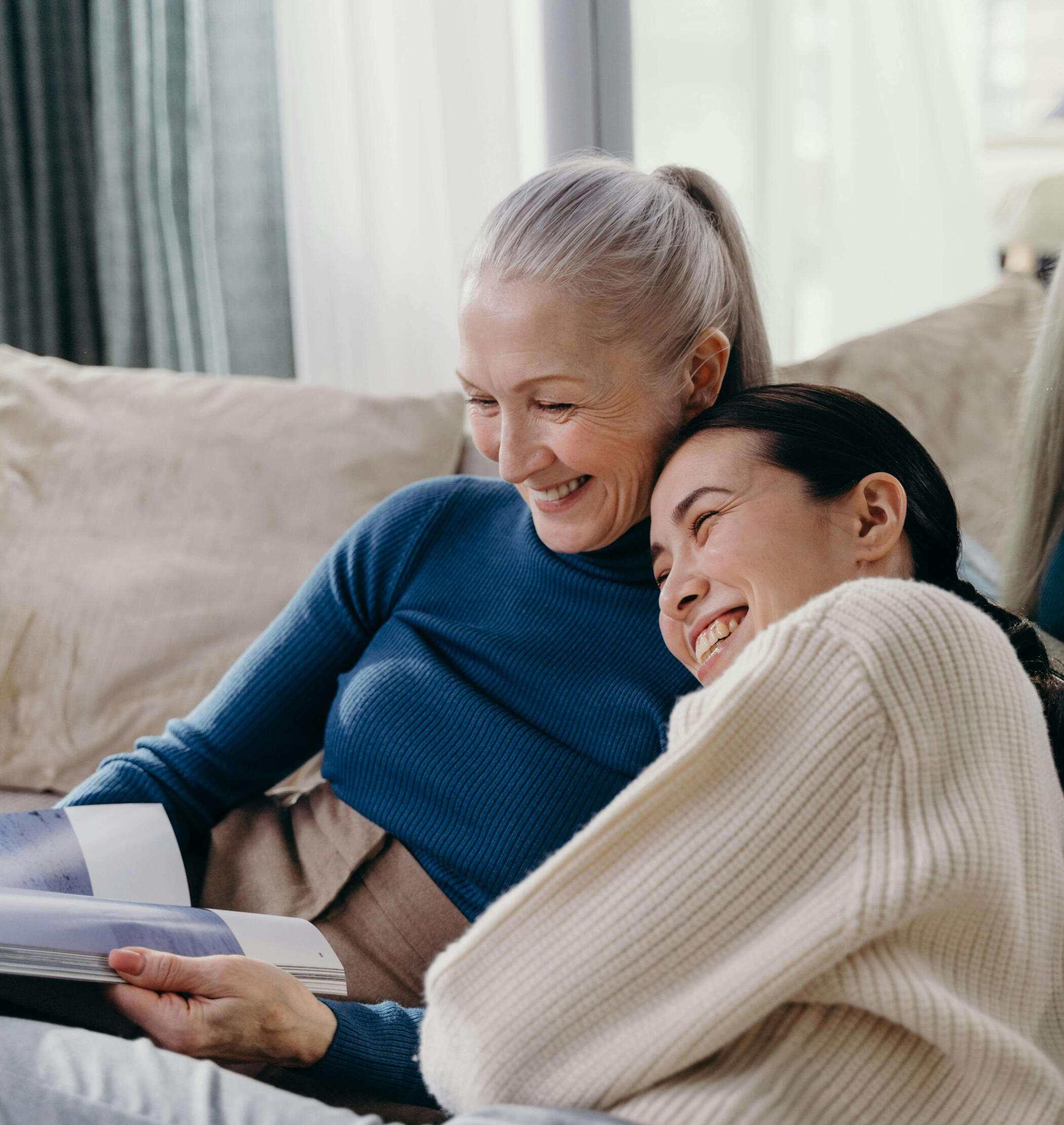 An older woman and a younger woman sit closely together on a couch, smiling as they read a document. Their relaxed posture and shared attention suggest warmth, support, and preparing for something important, such as a health visit.