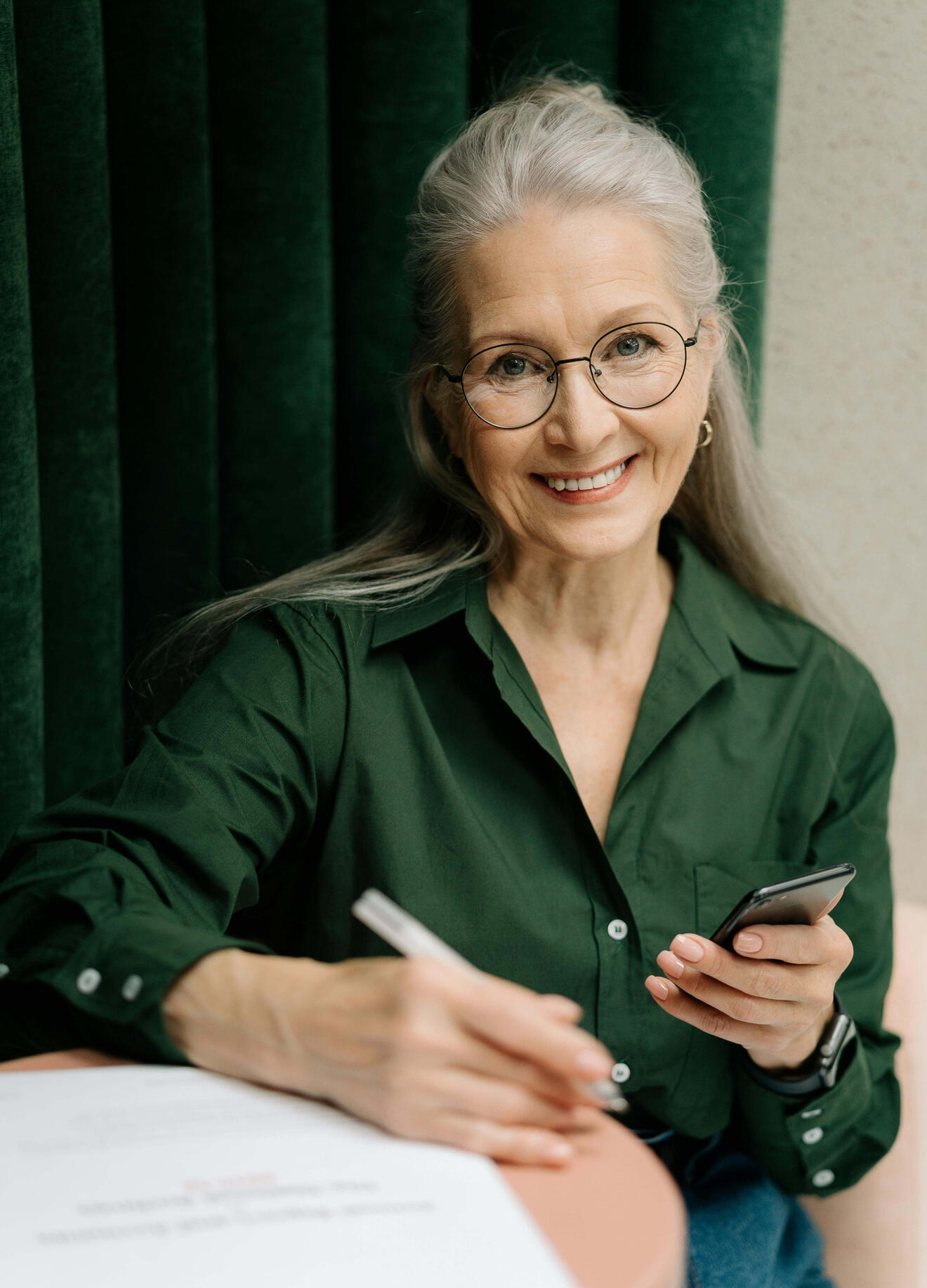 A smiling older woman sits at a kitchen table holding a smartphone in one hand and a printed health document in the other. The setting is calm and homely, suggesting comfort with both digital and printed information.